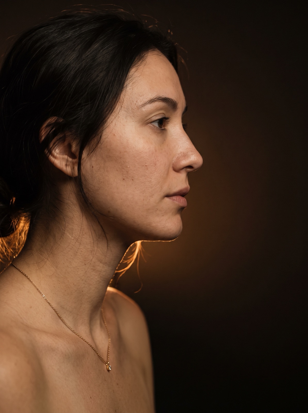 A woman in profile in low warm light, wearing a single delicate gold necklace